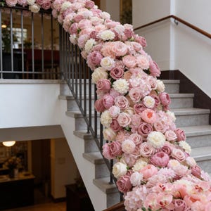 May include: A staircase railing adorned with a cascade of pink and white flowers. The arrangement features roses, peonies, and other blooms in varying shades of pink, creating a romantic and elegant display. The flowers are arranged to flow down the staircase.