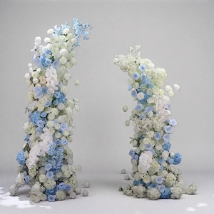 Op de afbeelding: Twee bloemenbogen bestaande uit witte en lichtblauwe bloemen. De bloemen zijn gerangschikt in een trapsgewijs ontwerp, waardoor een visueel aantrekkelijke weergave ontstaat. De bogen staan tegen een effen witte achtergrond, waardoor de bloemstukken worden benadrukt.