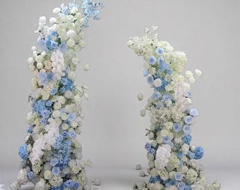 Puede incluir: Dos arcos florales compuestos por flores blancas y azul claro. Las flores están dispuestas en un diseño en cascada, creando una exhibición visualmente atractiva. Los arcos se colocan sobre un fondo blanco liso, resaltando los arreglos florales.
