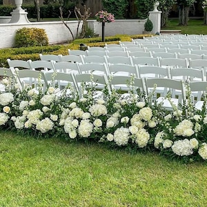 May include: A wedding ceremony setup with rows of white folding chairs. White flowers and greenery are arranged in front of the chairs. The scene is set on a green lawn with a white wall and trees in the background.