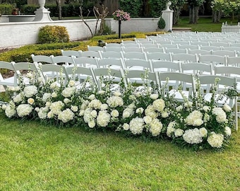 以下が含まれることがあります： 白い折りたたみ椅子が並んだ屋外の結婚式のセットアップ。白い花と緑が椅子の前に並んでいます。シーンは、白い壁と背景に木々がある緑の芝生に設定されています。