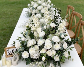 May include: A table setting with a white tablecloth, decorated with abundant white roses, snapdragons, and foliage. Wooden chairs and candles are placed near the floral arrangement. A wooden calendar shows "09 OCT".