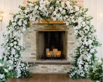 May include: A white floral archway decorates a brick fireplace with a lit candle arrangement inside. The archway is made of white flowers and greenery.