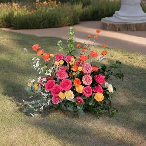 May include: A vibrant floral arrangement featuring a mix of pink, orange, and yellow roses, dahlias, and other flowers. The bouquet is set on a bed of green grass, with a blurred background of a garden and a white pillar.