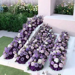 Puede incluir: Un arreglo floral con una escalera adornada con rosas moradas, lavanda y blancas. Las flores caen en cascada por los escalones, creando una exhibición visualmente impresionante. El arreglo se encuentra sobre un fondo de vegetación y una pared rosa claro.