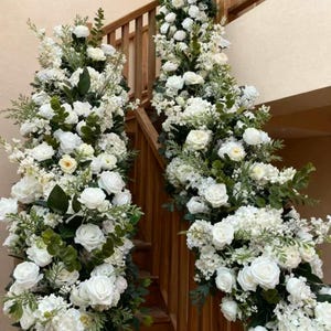 May include: Two staircases decorated with white roses, hydrangeas, and greenery. The floral arrangements cascade down the wooden stairs and along the handrails. The flowers are a mix of sizes and textures, creating a lush, elegant display.