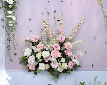 May include: A display of flowers, including pink and white roses and carnations, against a light purple backdrop. Green leaves and stems provide contrast. The arrangement is full and lush, with the flowers clustered together. The overall effect is soft and romantic.