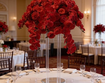 May include: A striking table centrepiece composed of rich red roses and orchids. The floral arrangement is supported by a clear, cylindrical stand, creating an elegant focal point for a formal event. The table is set with glassware and cutlery.
