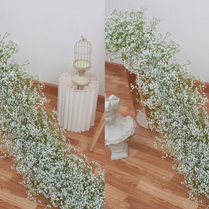 May include: Two white baby's breath garlands draped over gold metal stands. The garlands are made of small white flowers and green stems. The stands are on a wooden floor.