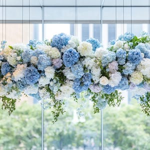 Pode incluir: Um arranjo floral suspenso, com hortênsias azuis, brancas e roxas claras. A folhagem verde cai, criando uma exibição natural e elegante. As flores estão em frente a uma grande janela.