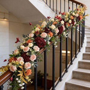 May include: A staircase with a floral garland. The garland features roses, hydrangeas, and other flowers in shades of pink, orange, burgundy, and cream. The staircase has a dark wooden handrail and black metal balusters.