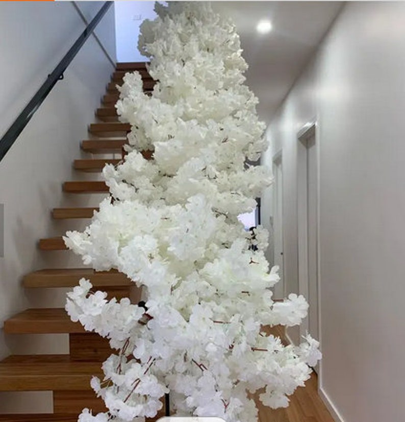 May include: A large, artificial white cherry blossom tree. The tree is positioned next to a wooden staircase with a black handrail. The blossoms are dense and create a full, textured appearance. The background includes a hallway and white walls.