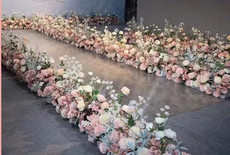 May include: A floral arrangement featuring rows of pink and white flowers, creating a pathway effect. The flowers are arranged in rectangular containers, with a variety of blooms and greenery. The overall color palette is soft and romantic, suitable for weddings or events.