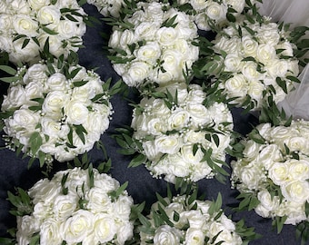 May include: Ten white silk rose bouquets with green leaves and white baby's breath. The bouquets are arranged in a circle on a dark gray surface.