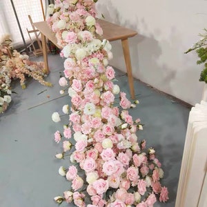May include: A long garland of pink and white artificial flowers, cascading down from a wooden table. The garland is made up of roses, carnations, and other floral elements.