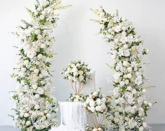 May include: Two white flower arrangements with greenery, each standing tall on a white base. The arrangements are made of white flowers and greenery, and are placed on a white surface.