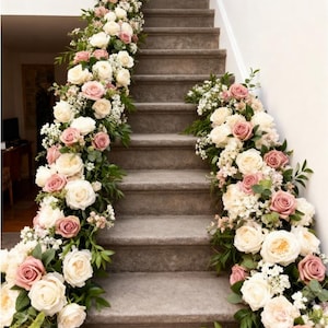 May include: A staircase decorated with floral arrangements. The steps are adorned with garlands of white and dusty rose roses, accented by greenery and small white flowers. The arrangement cascades down the side of the stairs.