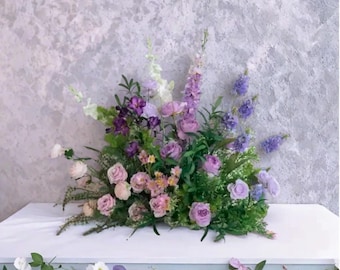 Könnte beinhalten: Ein Blumenarrangement mit lila, rosa und weißen Blumen, darunter Rosen und Lavendel. Die Anordnung befindet sich auf einer weißen Oberfläche mit einem strukturierten grauen Hintergrund. Die Blumen werden durch grünes Laub ergänzt.