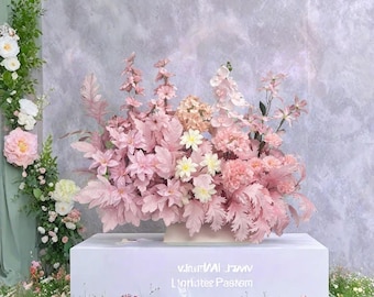 May include: A large floral arrangement in shades of pink, including dahlias and other blooms, displayed on a white pedestal. The arrangement is accented with small white flowers and greenery. The text "V.InutWAI J.MV L'oriatee Pastem" is visible on the pedestal.