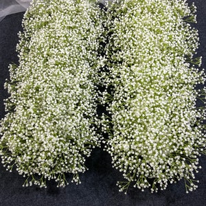 Puede incluir: Dos arreglos florales largos y blancos hechos de gypsophila, dispuestos simétricamente sobre una superficie gris oscuro.