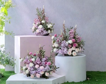 Könnte beinhalten: Drei Blumenarrangements mit weißen, rosa und lila Blumen. Die Arrangements stehen auf weißen und rosa geometrischen Podesten vor einem hellgrauen Hintergrund und grünem Gras. Die Blumen umfassen Rosen, Hortensien und andere Blüten.
