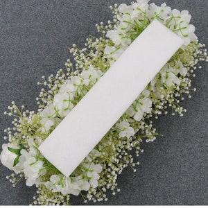 May include: A rectangular white floral foam block surrounded by white flowers and small white buds. The arrangement is set against a gray background, creating a contrast that highlights the floral display. The flowers appear to be artificial.