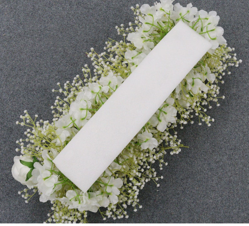 May include: A rectangular white floral foam block surrounded by white flowers and small white buds. The arrangement is set against a gray background, creating a contrast that highlights the floral display. The flowers appear to be artificial.