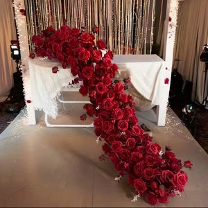 May include: A long garland of deep red artificial roses cascading down a white table. The roses are arranged in a flowing, natural pattern, creating a romantic and elegant backdrop.