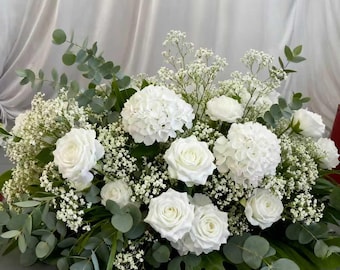 May include: A low floral arrangement of white roses, hydrangeas, and baby's breath, with green eucalyptus foliage. The flowers are arranged in a rectangular shape, suitable for a table centrepiece or other decorative use.