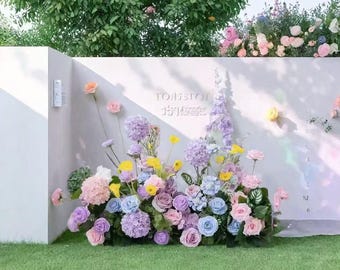 Könnte beinhalten: Ein Blumenarrangement mit pastellfarbenen Blumen, darunter Rosen, Hortensien und andere Blüten. Die Blumen sind vor einer weißen Wand mit Text angeordnet. Das Arrangement steht auf einer grünen Wiese.