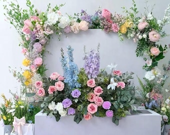 Könnte beinhalten: Ein lebendiges Blumenarrangement mit einer Vielzahl farbenfroher Blumen, darunter rosa Rosen, blaue Rittersporne und weiße Lilien. Die Blumen sind in einem rechteckigen Rahmen und auf einer weißen Oberfläche angeordnet.