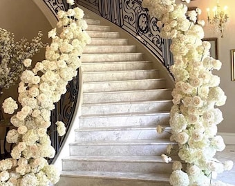 May include: A white marble staircase with a black wrought iron railing is decorated with white flowers. The flowers are arranged in a cascading design on either side of the staircase.