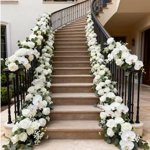 May include: A curved staircase adorned with white roses and orchids, creating a floral garland along the black iron railings. The steps are a light beige, and the scene is bathed in natural light.