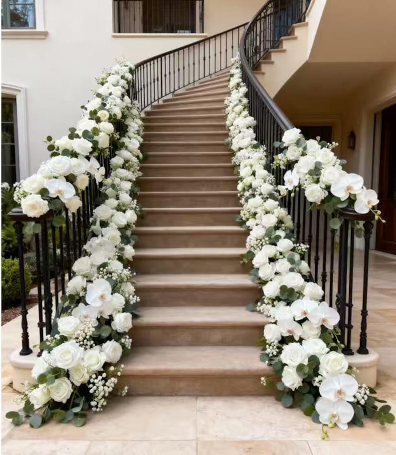 May include: A curved staircase adorned with white roses and orchids, creating a floral garland along the black iron railings. The steps are a light beige, and the scene is bathed in natural light.