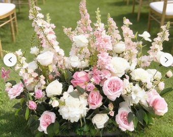 May include: A floral arrangement with pink and white roses, hydrangeas, and other flowers. The arrangement is set on a bed of green leaves and is placed on a grassy lawn. Gold chairs and a white table are in the background, suggesting a wedding or event setting.