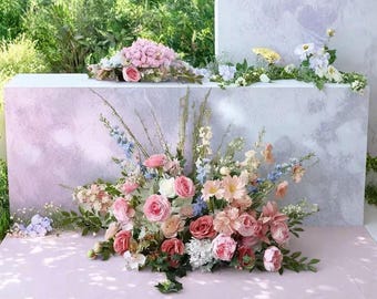 May include: A display of mixed flowers, including pink and red roses, with blue and peach accents. The arrangement is set against a backdrop of light pink and grey, with green foliage.