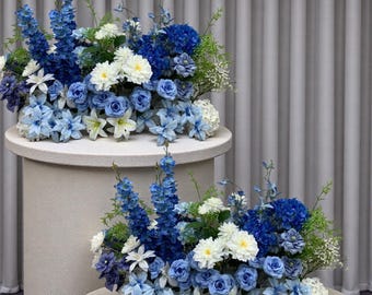 Puede incluir: Dos arreglos florales con flores azules y blancas, incluyendo hortensias, rosas y lirios. Los arreglos se exhiben sobre pedestales cilíndricos blancos, contra un fondo neutro. Las flores varían en tonos de azul claro a oscuro.