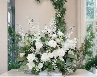 Op de afbeelding: Een weelderig bloemstuk met witte rozen, leeuwenbekjes en gipskruid. De bloemen worden geaccentueerd met groen blad en gepresenteerd op een marmeren oppervlak. Ideaal voor bruiloften of speciale evenementen.