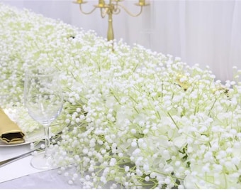 May include: A white baby's breath garland, also known as gypsophila, is draped across a table setting. The garland is made of small white flowers and green stems. A clear glass wine glass is on the table, along with a gold napkin.