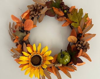 May include: A fall-themed wreath with a sunflower, pine cones, and leaves in shades of brown, orange, and green. The wreath is made of twigs and has a rustic look.