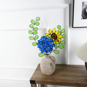 May include: A decorative vase holds a colorful arrangement of faux flowers. The arrangement includes a blue hydrangea, a yellow sunflower, and green leaves. The vase is a light beige color and sits on a wooden table.