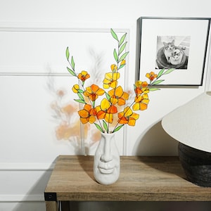 May include: A white ceramic vase with a face design holds a bouquet of orange and yellow flowers with green stems and leaves. The vase sits on a wooden table, with a framed black and white photo and a lamp in the background.