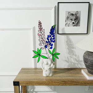 Può includere: Un arrangiamento floreale in vetro colorato con fiori di lupino rosa e blu con foglie verdi in un vaso di ceramica bianco a forma di busto femminile.