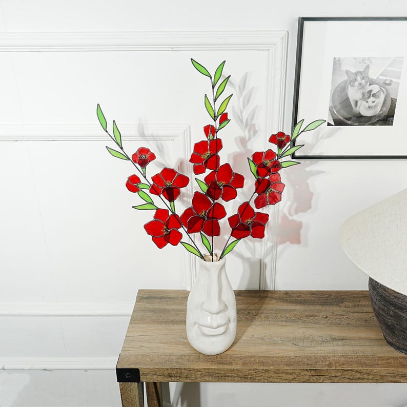 May include: A white ceramic vase with a face design holds a bouquet of red flowers with green stems and leaves. The vase sits on a wooden table, with a framed black and white photo in the background.