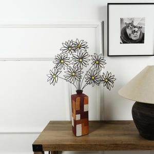 May include: A decorative metal daisy bouquet in a geometric patterned vase. The vase is brown, orange, and white. The daisies have white petals and yellow centers. The arrangement sits on a wooden table.
