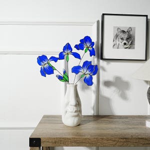 May include: A white ceramic vase with a face design holds several blue iris flowers with green stems. The flowers are a vibrant blue with white and yellow accents. A framed black and white photo of a dog hangs on the wall behind the vase.