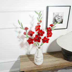 May include: A white ceramic vase with a face design holds red and green floral stems. The flowers have red petals and green leaves. The vase sits on a wooden table, with a framed black and white photo in the background.