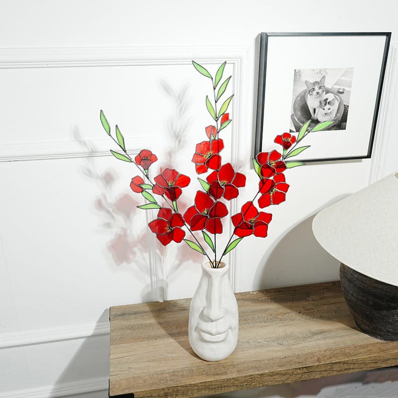 May include: A white ceramic vase with a face design holds red and green floral stems. The flowers have red petals and green leaves. The vase sits on a wooden table, with a framed black and white photo in the background.