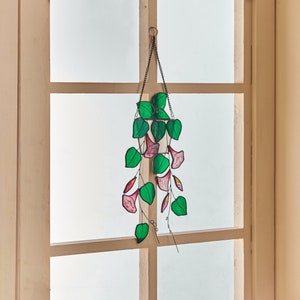 Peut inclure: Une suspension en verre color&eacute; repr&eacute;sentant une plante en pot avec des fleurs roses et des feuilles vertes. La plante est suspendue &agrave; une cha&icirc;ne noire.