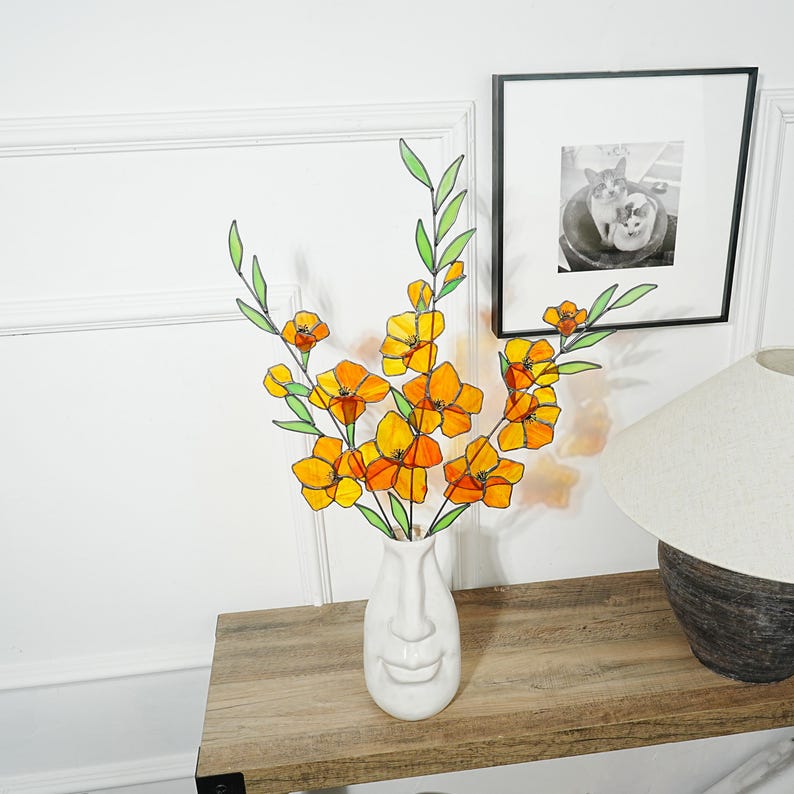 May include: A white ceramic vase with a face design holds a bouquet of orange and yellow flowers with green stems and leaves. The flowers are arranged on a wooden table, with a framed black and white photo in the background.